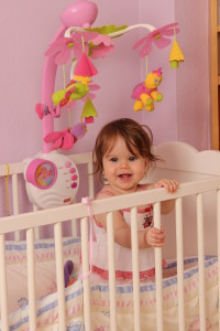 Маленькая девочка улыбается, стоит в своей кроватке - фото