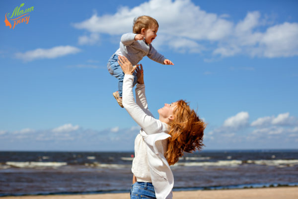 Redheaded mom in jeans and a white sweater throws up his young son against the sea and clouds